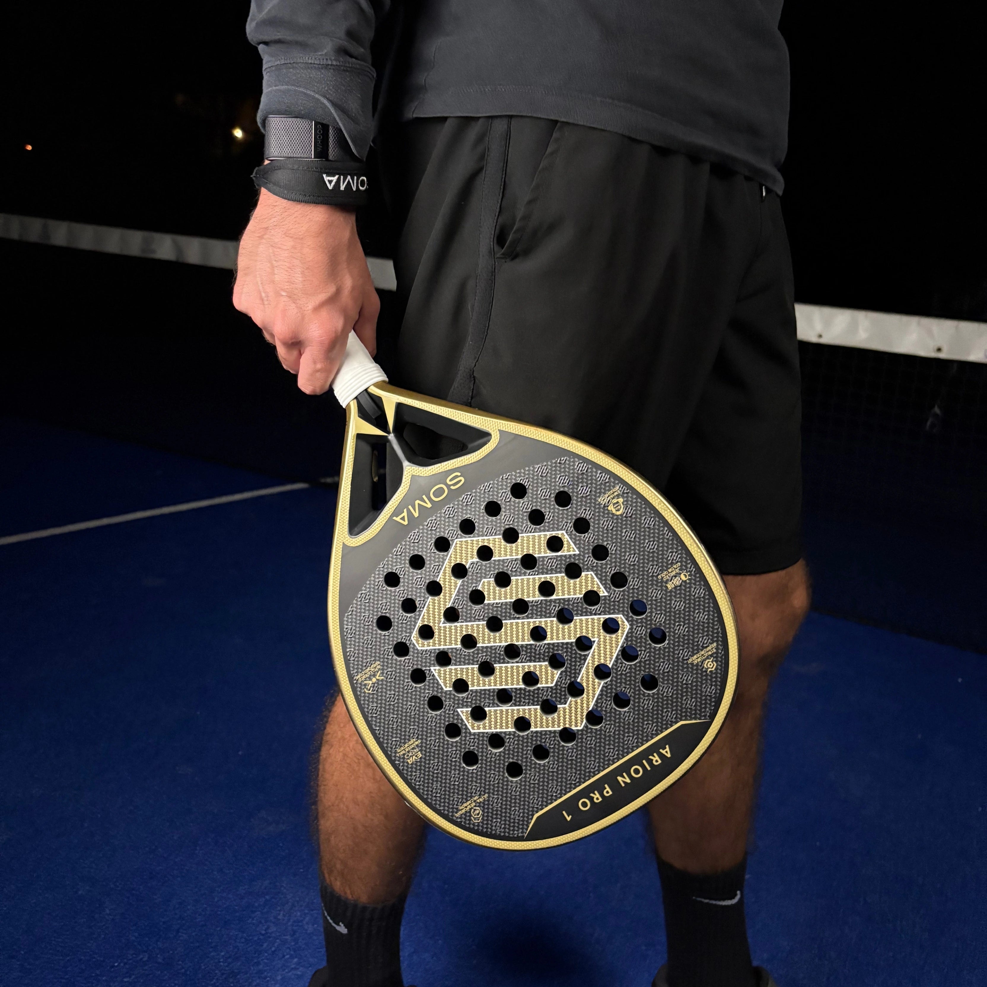 Raquette de padel Soma Arion Pro 1 tenue par un joueur sur terrain bleu- vue lifestyle avec poignet SOMA